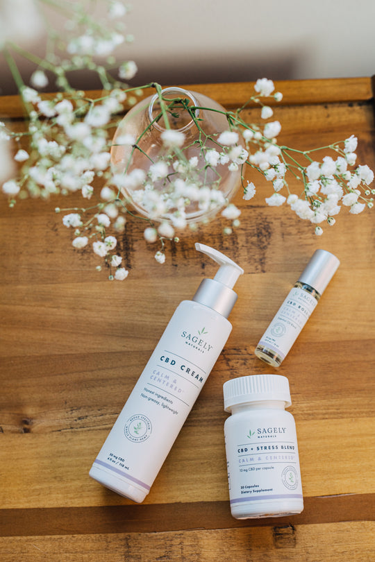 Sagely Naturals Calm & Centered CBD cream, CBD roll-on and CBD capsules on top of a wooden table, next to a vase of baby's breath.