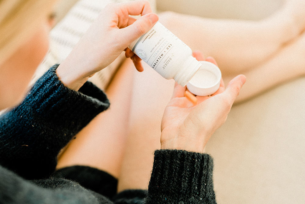 A woman holding Sagely Naturals Drift & Dream CBD + Melatonin capsules sitting on a tan sofa.