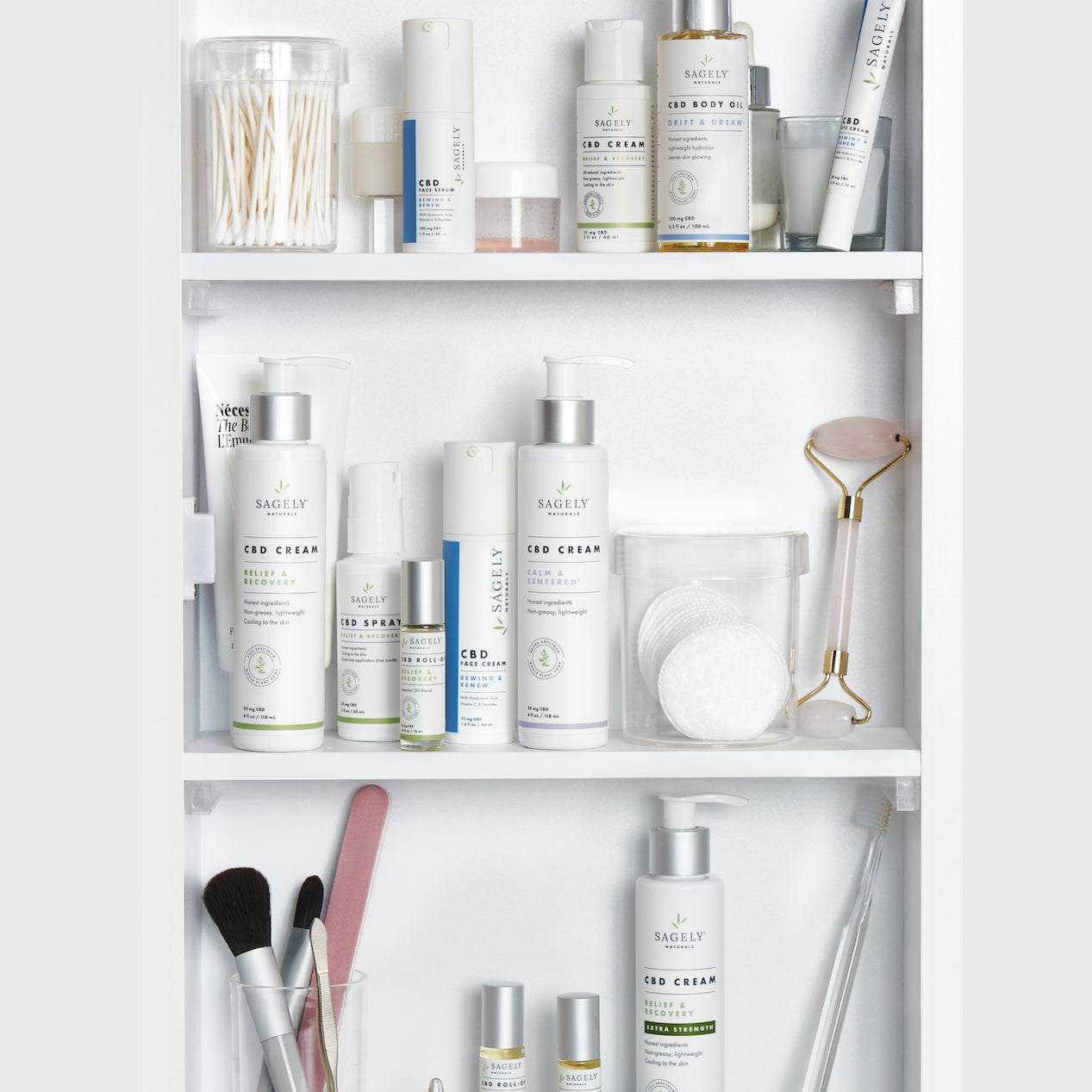 A medicine cabinet containing shelves with Sagely Naturals CBD cream, CBD capsules, CBD spray, CBD roll-ons, makeup brushes, makeup pads, and cotton buds.