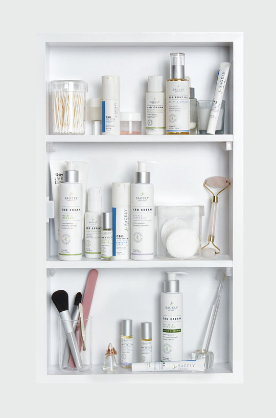 A medicine cabinet containing shelves with Sagely Naturals CBD cream, CBD capsules, CBD spray, CBD roll-ons, makeup brushes, makeup pads, and cotton buds.