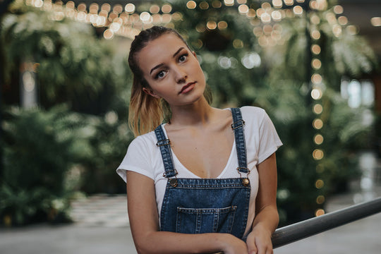 A young woman in denim overalls and a ponytail standing otuside