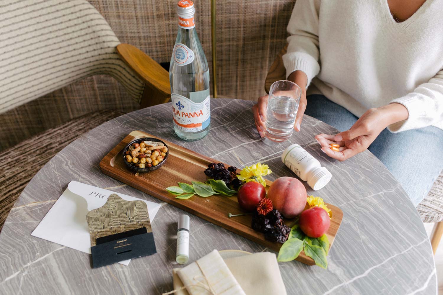 A woman taking Sagely Naturals CBD capsules while sitting at a table with fruit, nuts and sparkling water