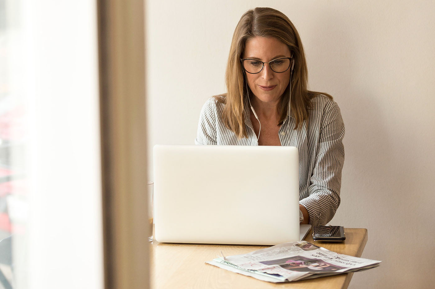 A woman siting at a laptop working with a newspaper and cellphone next to her