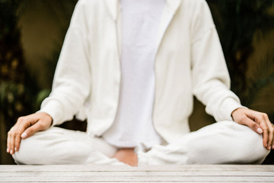 A woman in all white sitting cross-legged with her hands on her knees, practicing whole self wellness