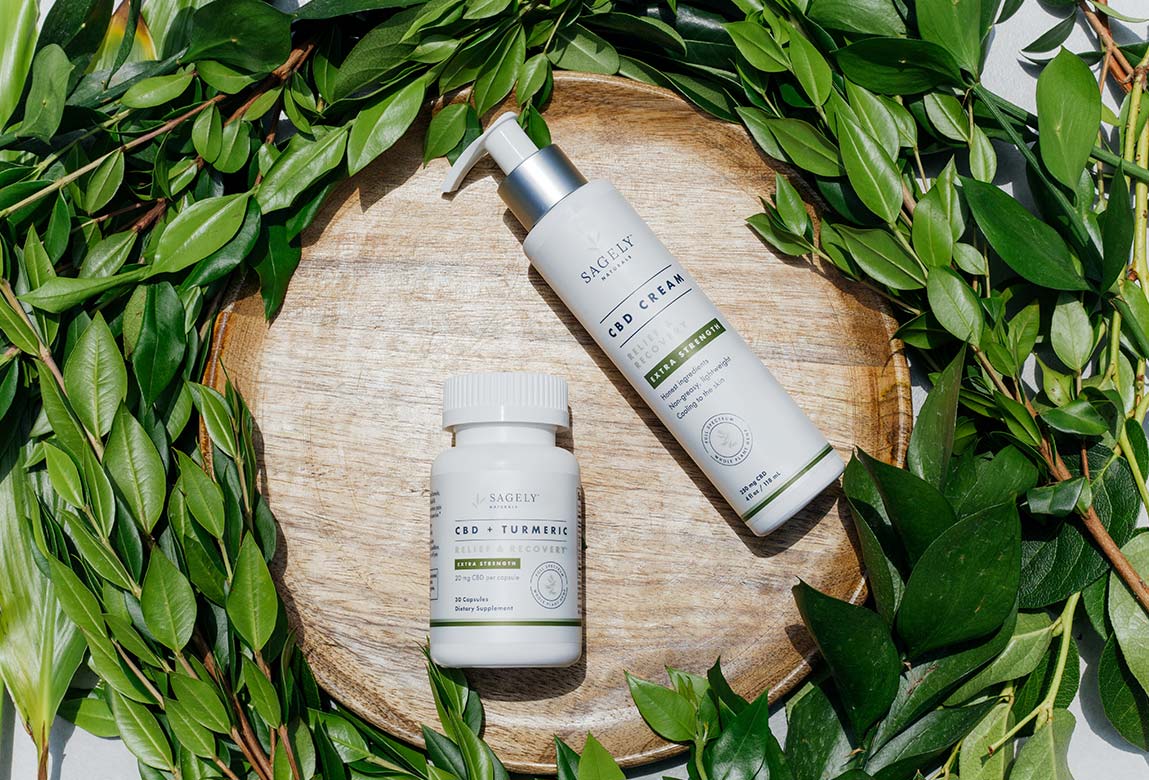 Sagely Naturals Relief & Recovery Extra Strength CBD Cream & CBD Capsules lying on a wooden plate surrounded by green leaves.