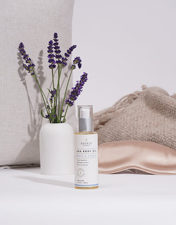 Sagely Naturals CBD Drift & Dream CBD Body Oil on bench in front of a lavender plant and eye mask.