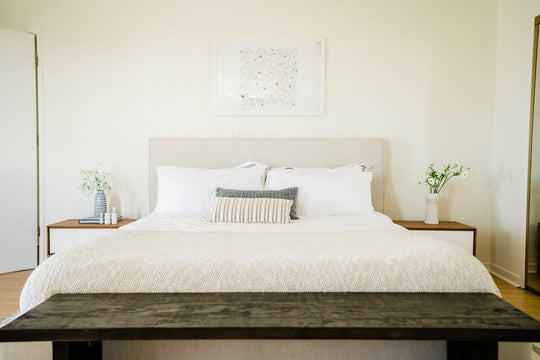 A bed with crisp white sheets, pillows, framed picture above bed, and bedside tables with flowers and Sagely Naturals CBD products on the bedside table.