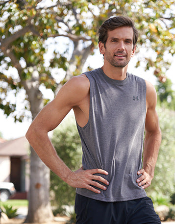 A man in gray workout singlet and shorts standing outdoors with his hands on hips.