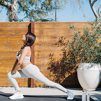 Woman with long brown hair stretching into a lunge stretch in all-white workout clothes in her outdoor space.