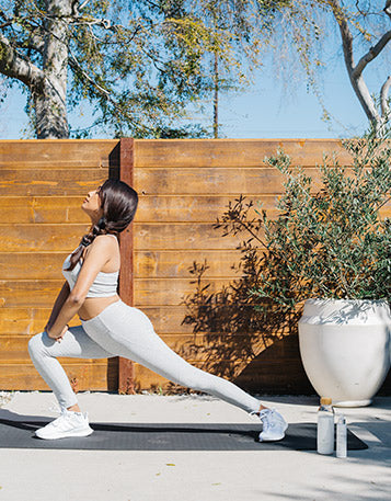 Woman with long brown hair stretching into a lunge stretch in all-white workout clothes in her outdoor space.