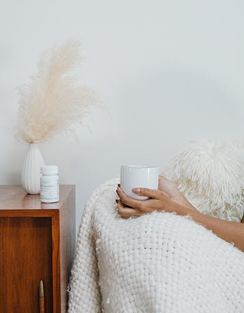 A woman’s hands holding a mug next to a cupboard with Sagely Naturals capsules and dried flower arrangement.