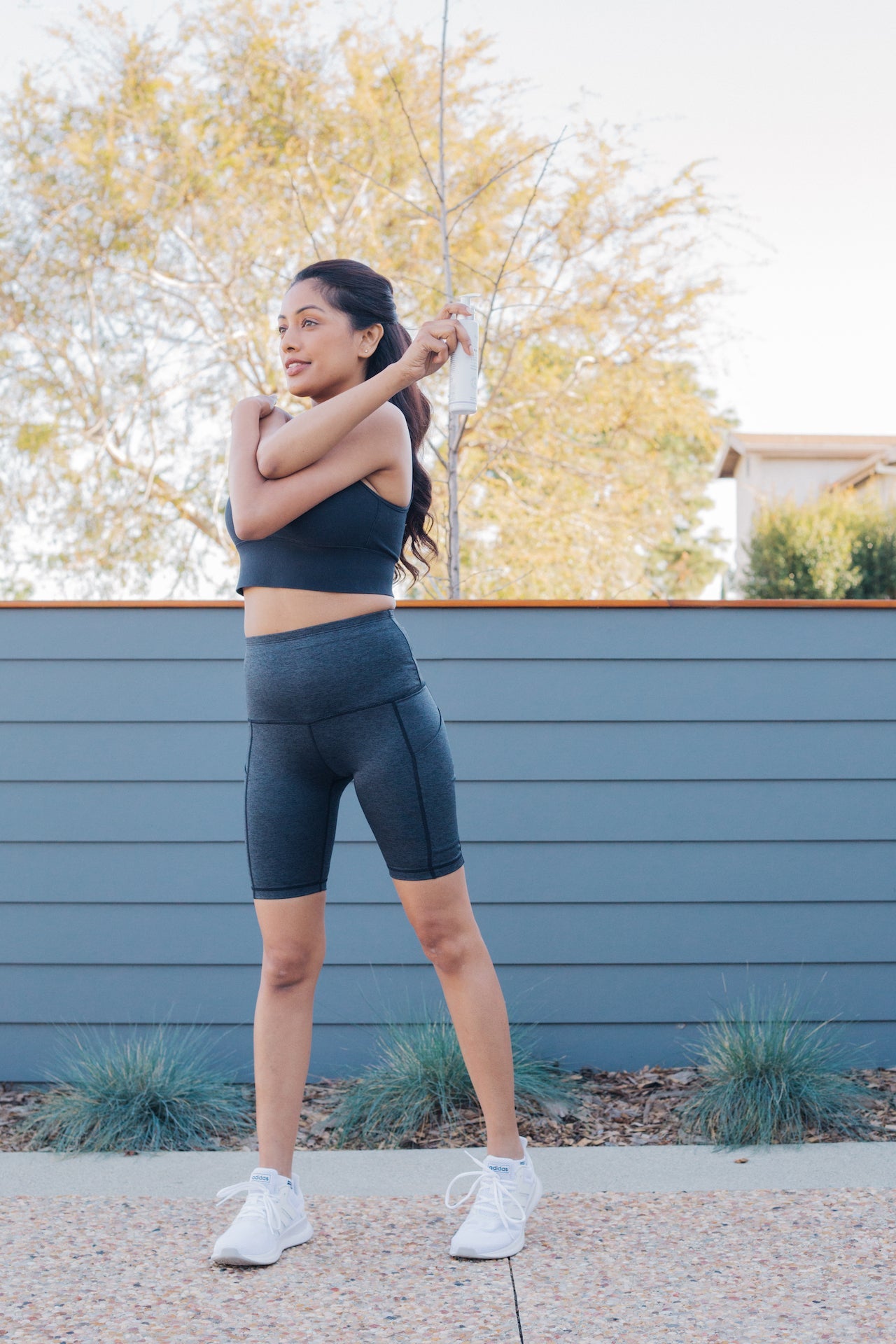 A woman in workout clothes standing outside doing a shoulder stretch while holding Sagely Naturals Relief & Recovery CBD cream