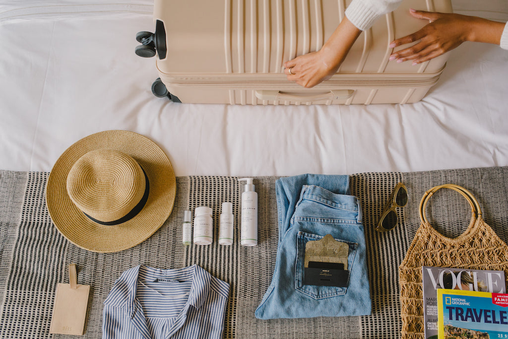 Sagely Naturals CBD products lying on bed next to suitcase, hat, sunglasses.